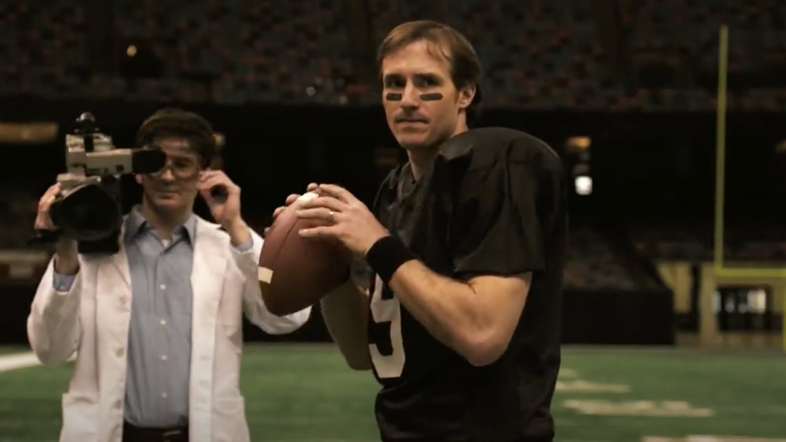 Drew Brees holds the football while a man in a white lab coat and a video camera stands next to him. 