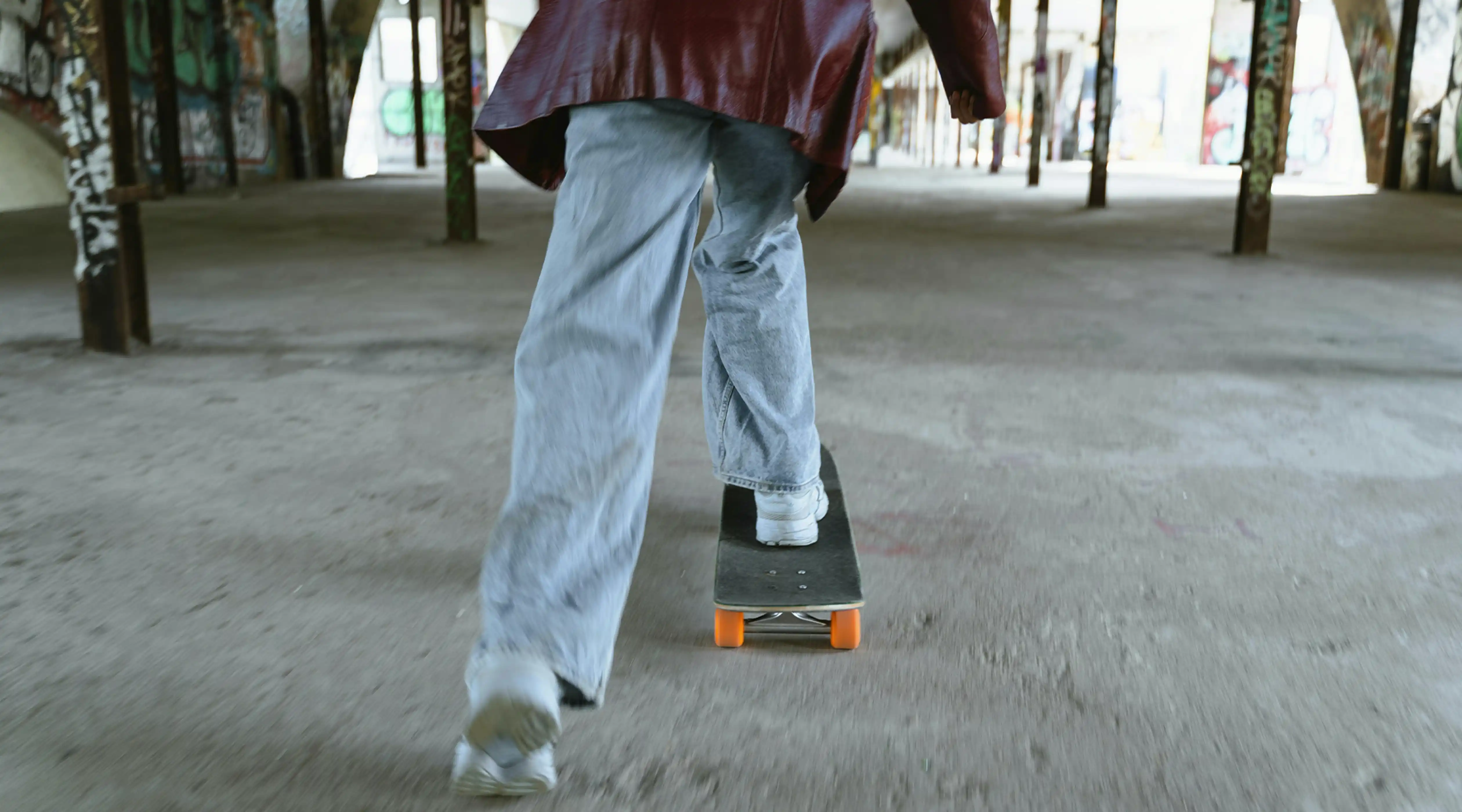 Person on a skateboard gliding away from camera through a graffiti-covered abandoned building with concrete pillars, wearing a maroon jacket and light jeans — representing the cultural momentum and forward motion at the heart of movement marketing strategy