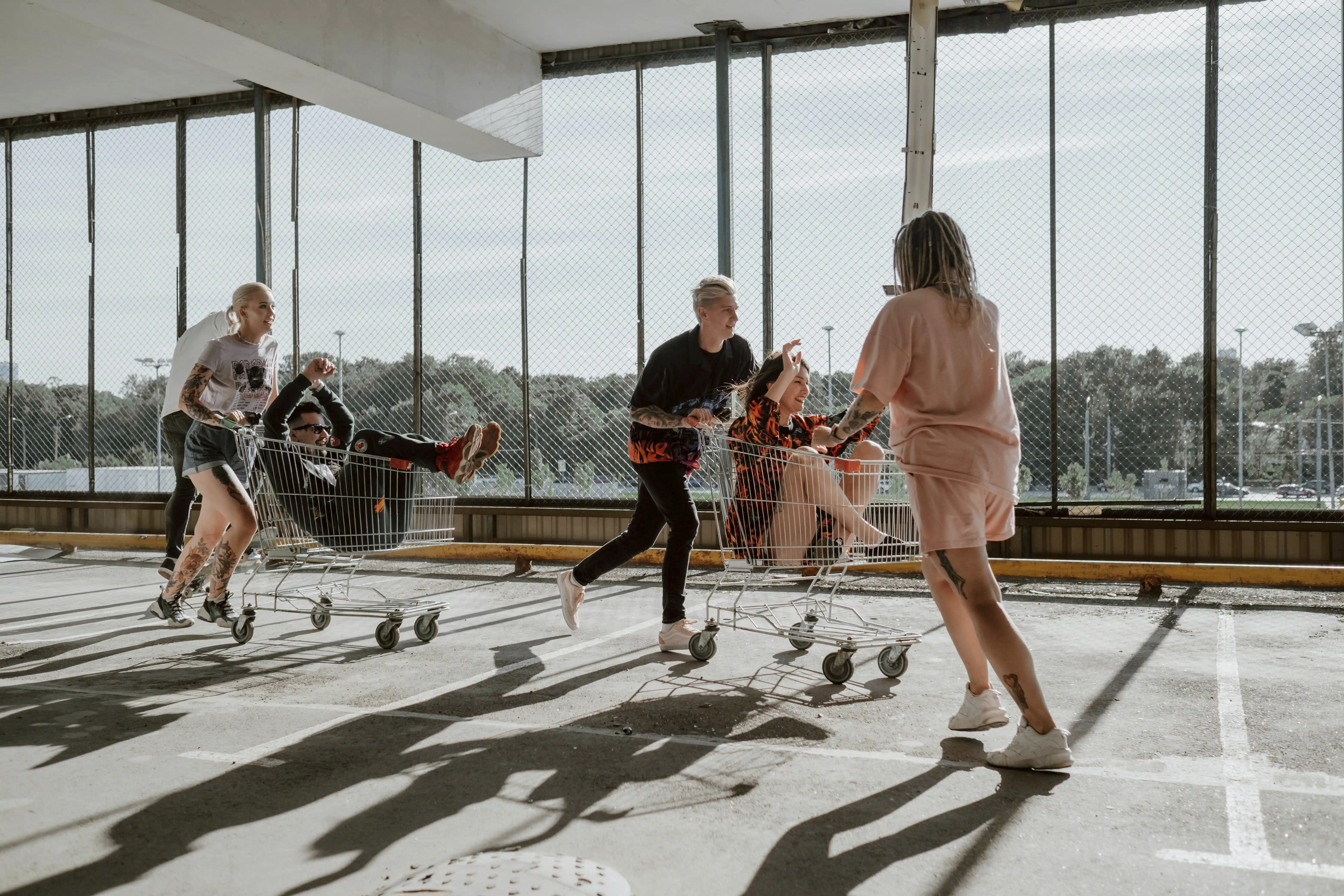 Group of young people laughing and racing each other in supermarket shopping carts through a sunlit parking garage with long dramatic shadows on the floor — capturing the spontaneous energy and community participation that defines movement marketing brand strategy