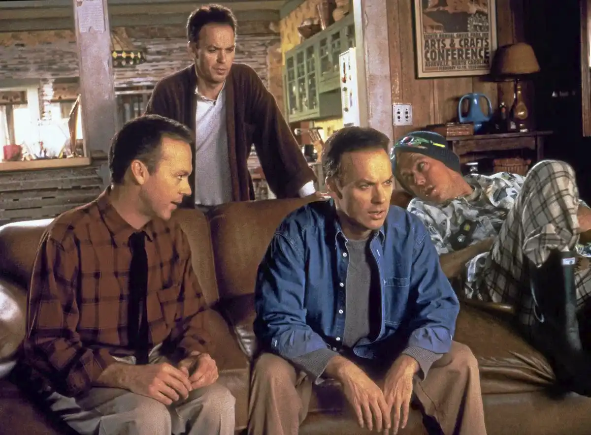 Film still from Multiplicity (1996) showing four versions of Michael Keaton's character Doug gathered in a rustic wood-paneled room — three engaged and attentive, one (Steve, the copy of a copy) lounging casually in the background, illustrating how each generation away from the original source loses something essential