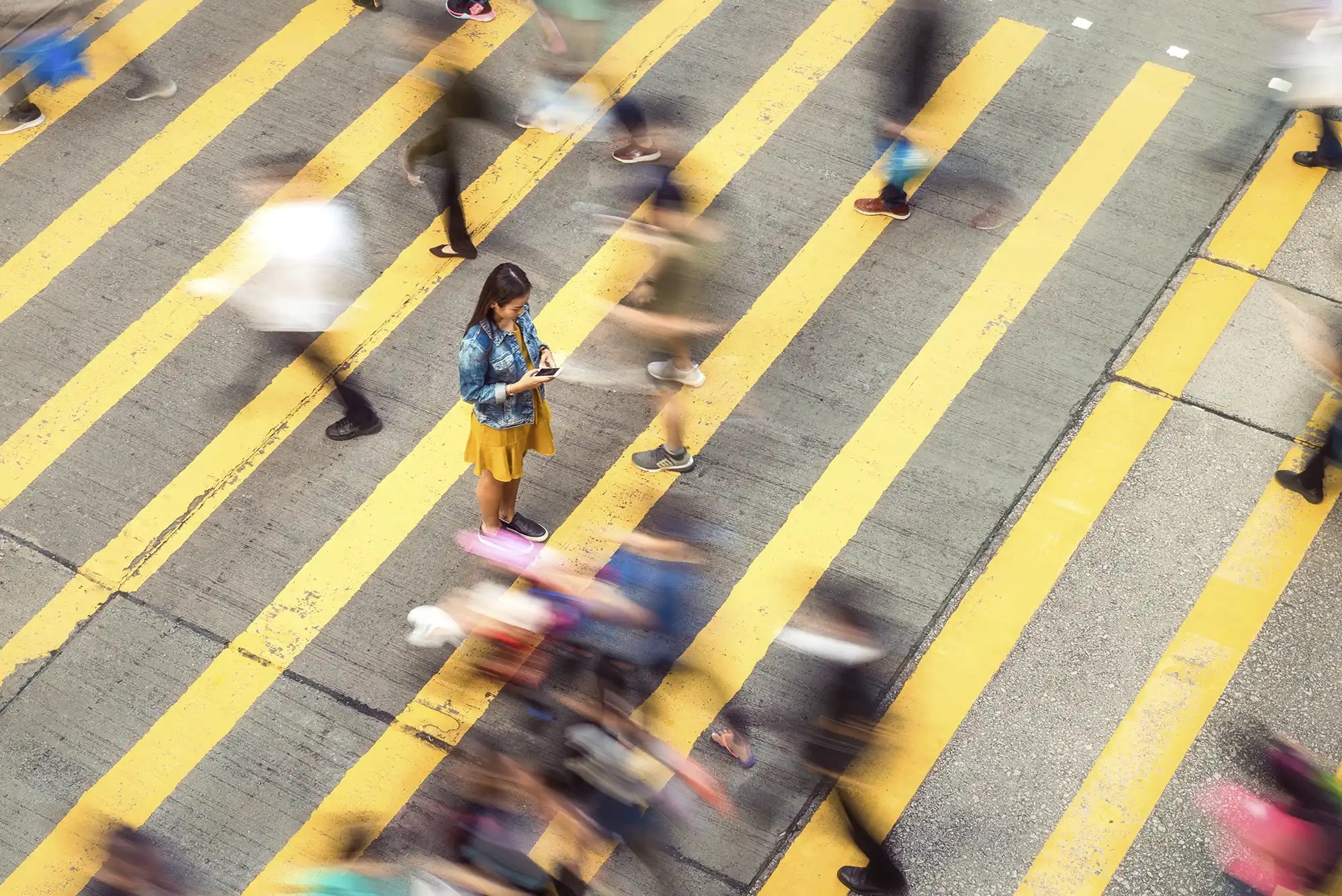 Woman in yellow dress standing still on a striped crosswalk, looking at her phone as blurred pedestrians rush past.