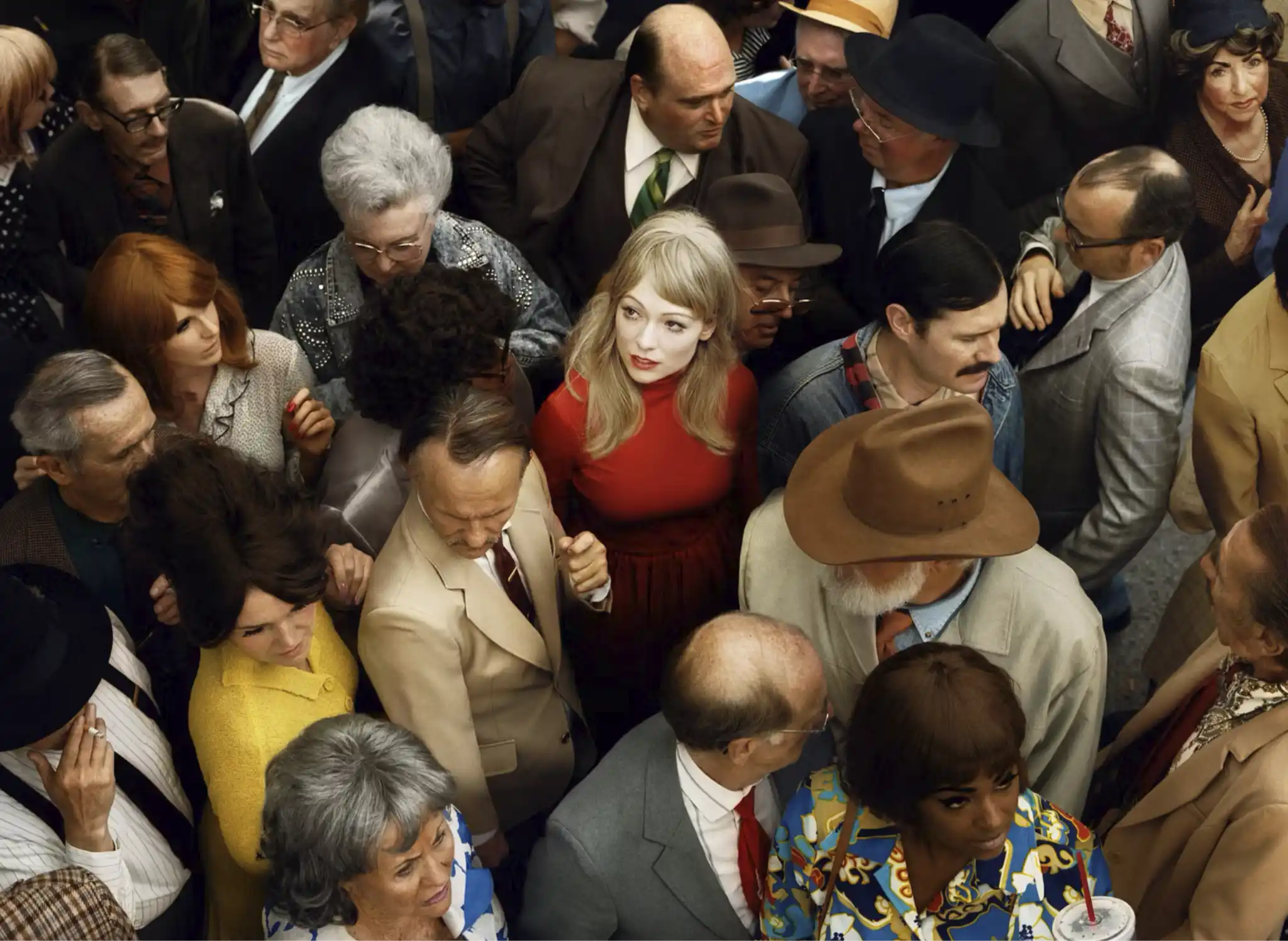 Overhead view of a crowded group of people in vintage-style clothing, with a blonde woman in a bright red dress at the center looking up amid the surrounding faces.