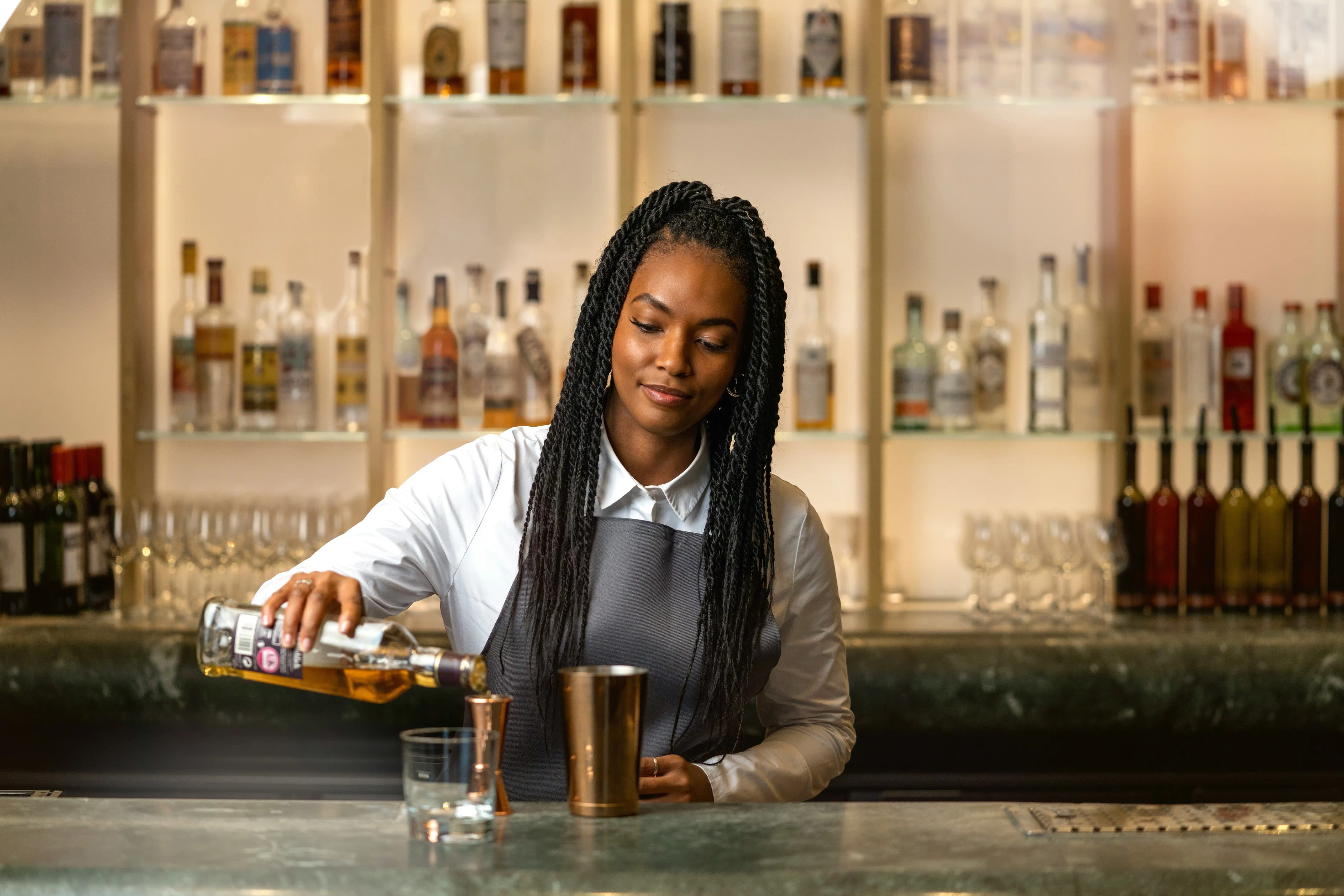 Female bartender pouring whiskey from a bottle into a jigger beside a metal shaker at a backlit bar, with shelves of bottles behind.