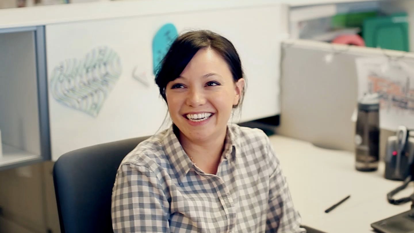 Woman wearing plaid with hair pulled back smiling with her mouth open at her work cubicle. Still from the “Momentum OnUp” campaign.
