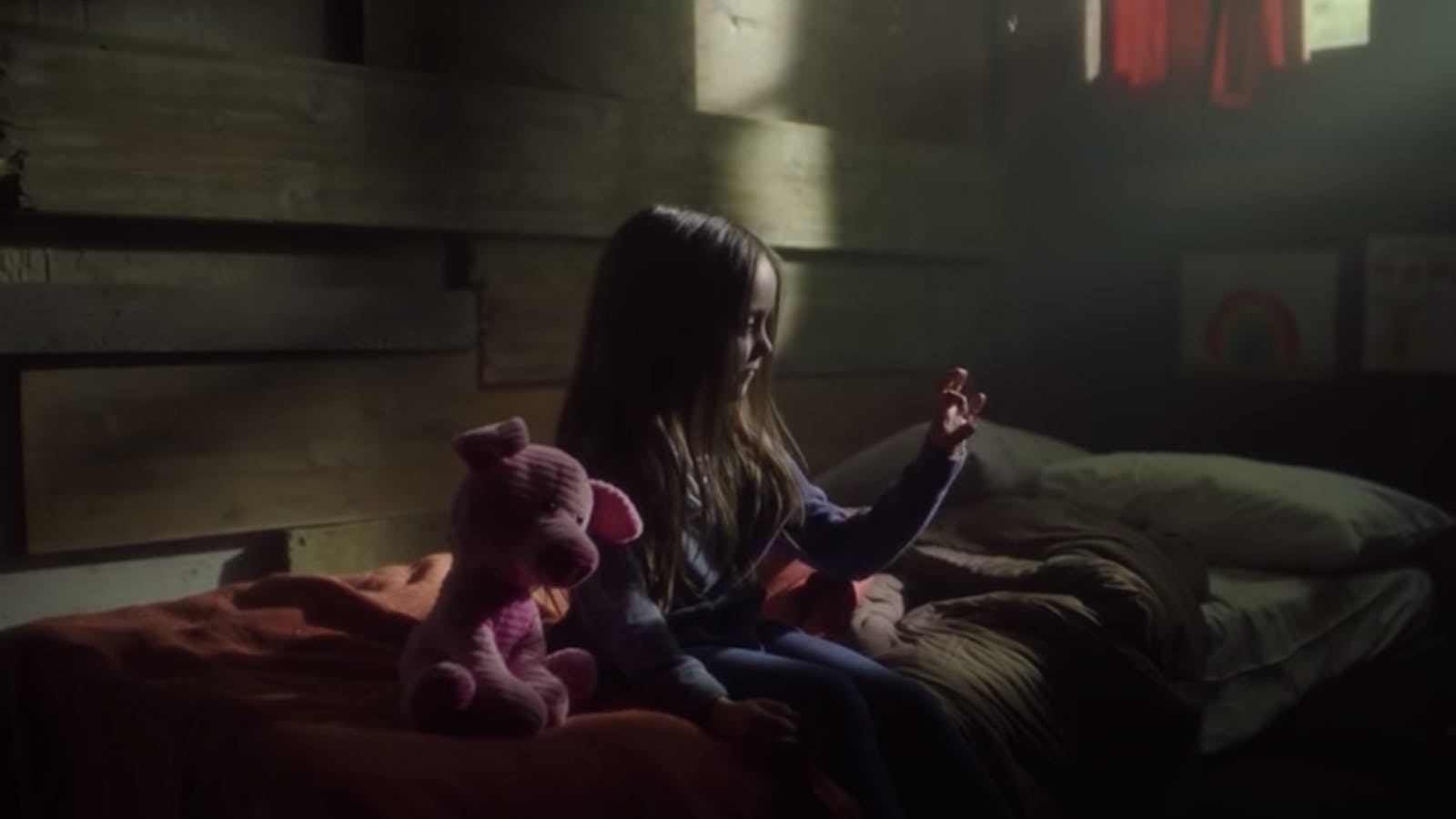 Screengrab of a young girl in a dark room, sitting on her bed next to her stuffed animal.
