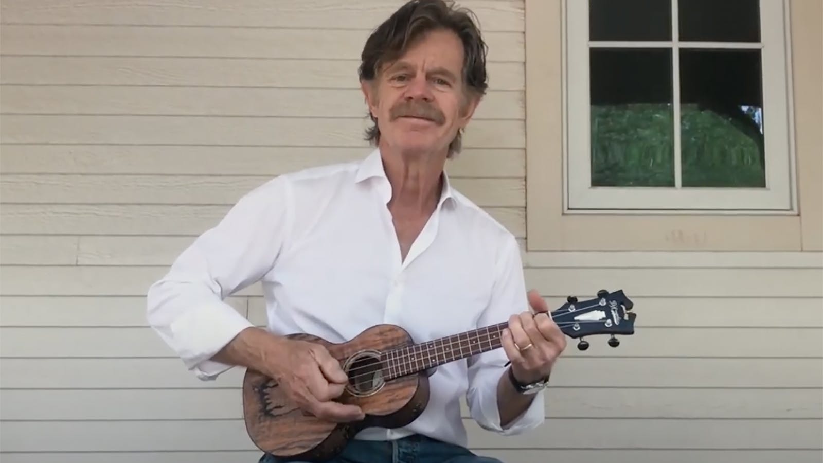 Actor William H. Macy wears a white button-up and plays a baby guitar in front of an off-white home.
