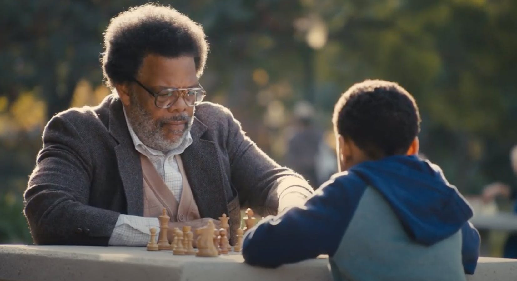 A middle aged bearded man with glasses plays chess outdoors with a boy.