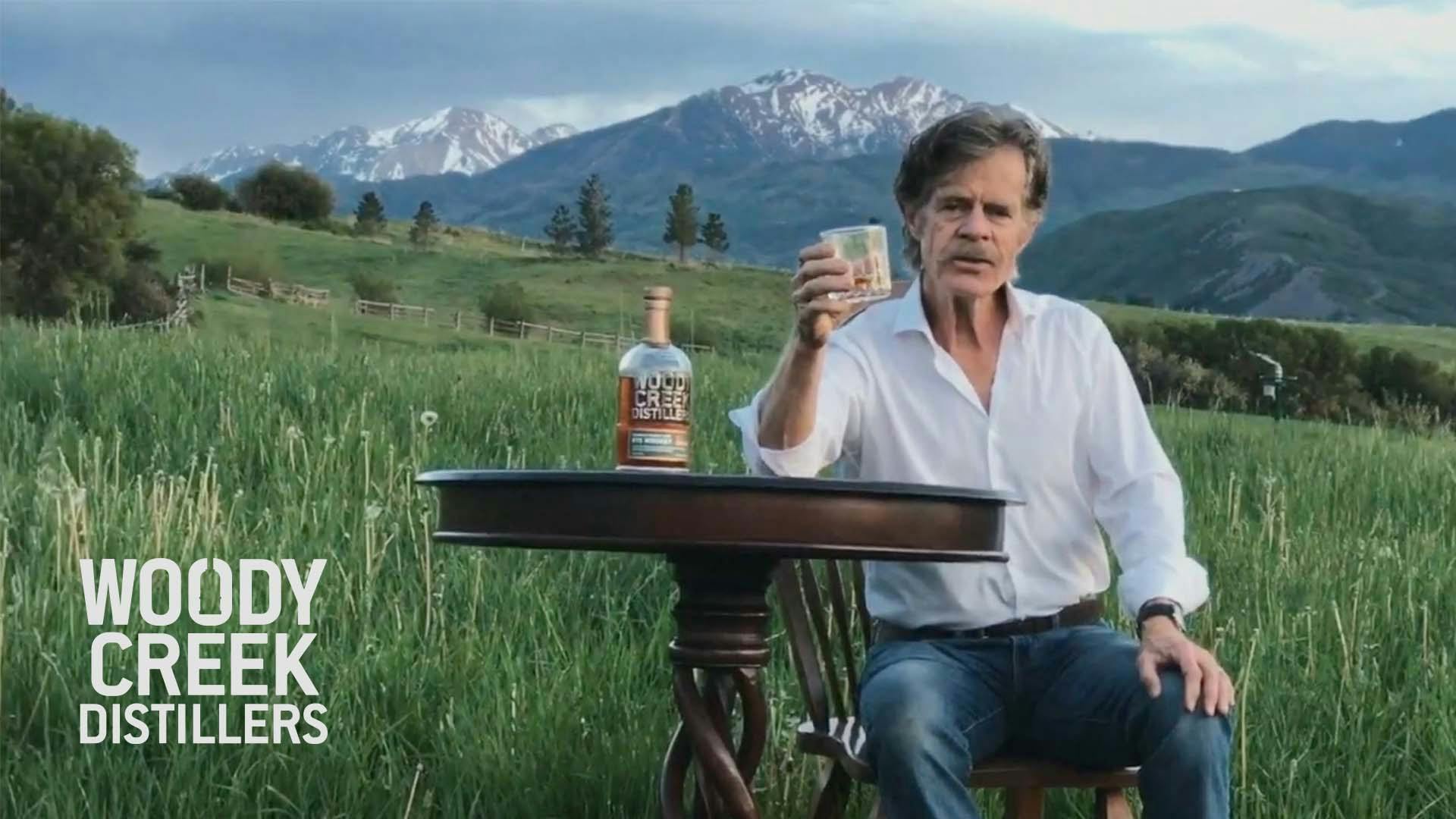 Actor William H. Macy holds a glass of Woody Creek bourbon on a field while wearing a white button-up.
