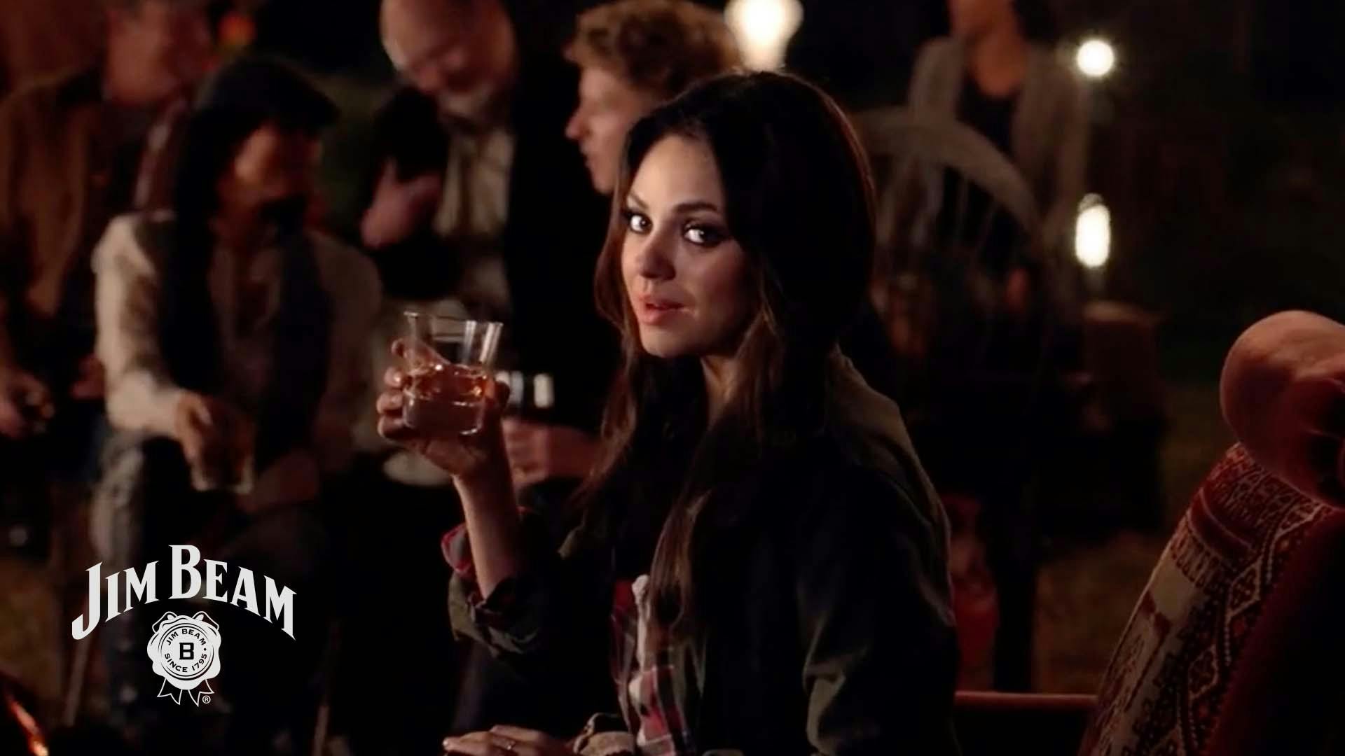 Actress Mila Kunis savors a glass of Jim Beam whiskey in a crowded room.