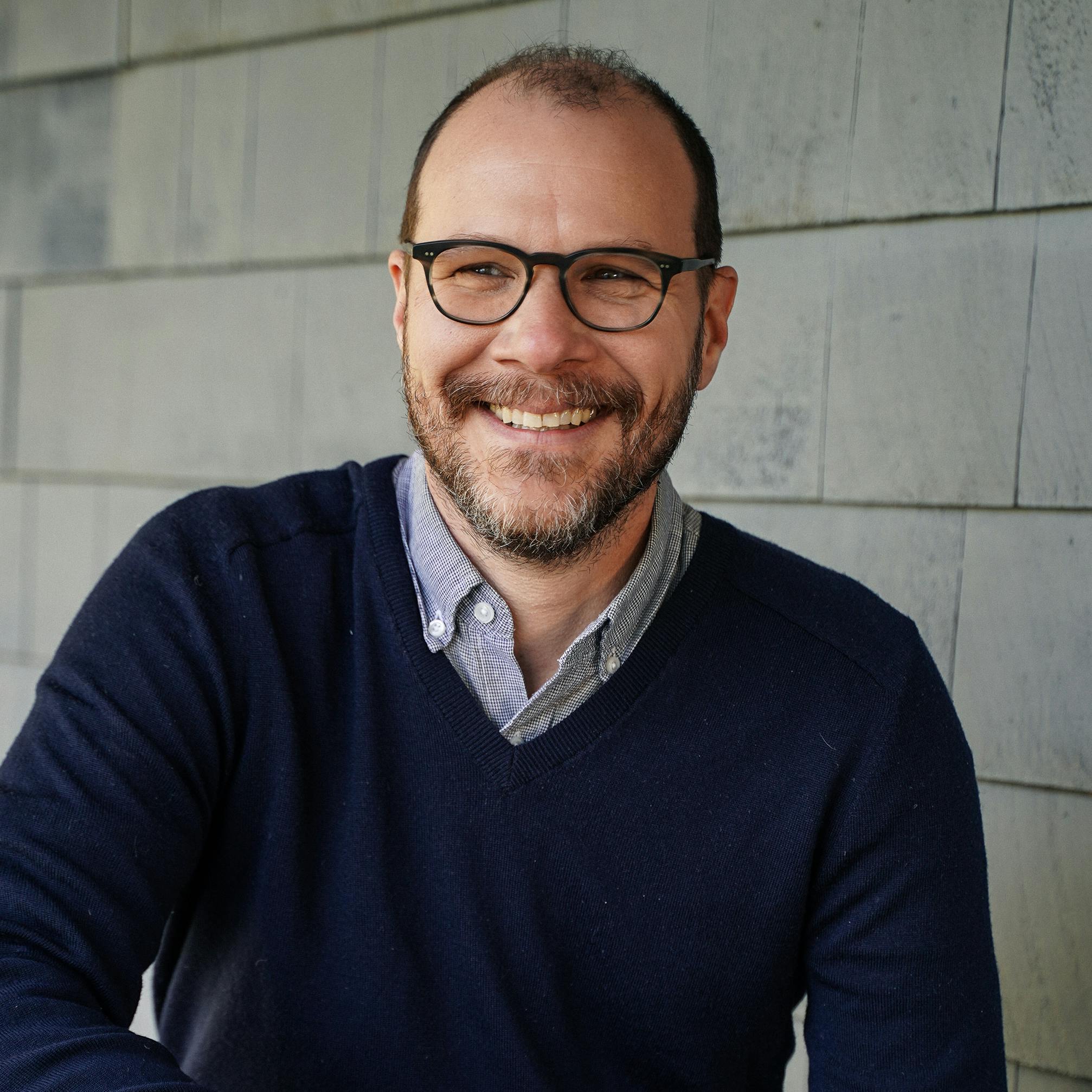 Portrait of StrawberryFrog Finance Director Jon Zeidner smiling in front of a gray wall. He is wearing glasses and a sweater with a button-down collar peeking through.