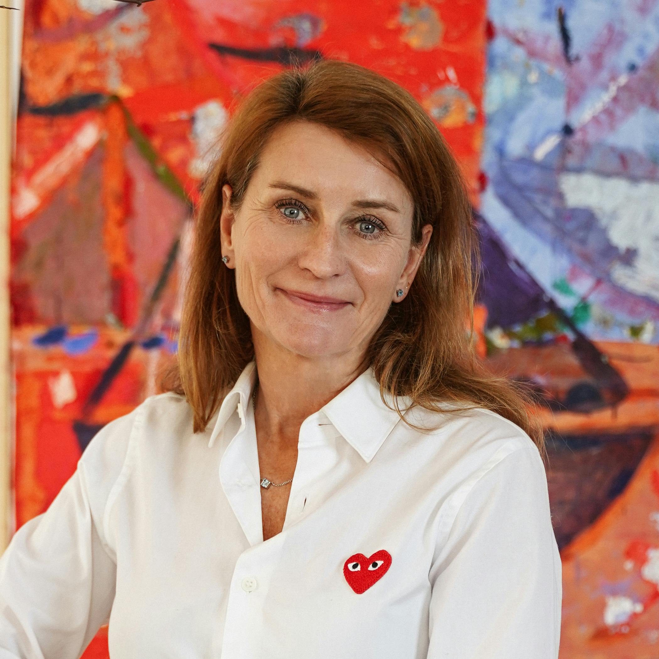 Portrait of StrawberryFrog Co-Founder & COO Karin Drakenberg wearing a white button-down, smiling softly in front of a brightly colored painting. 