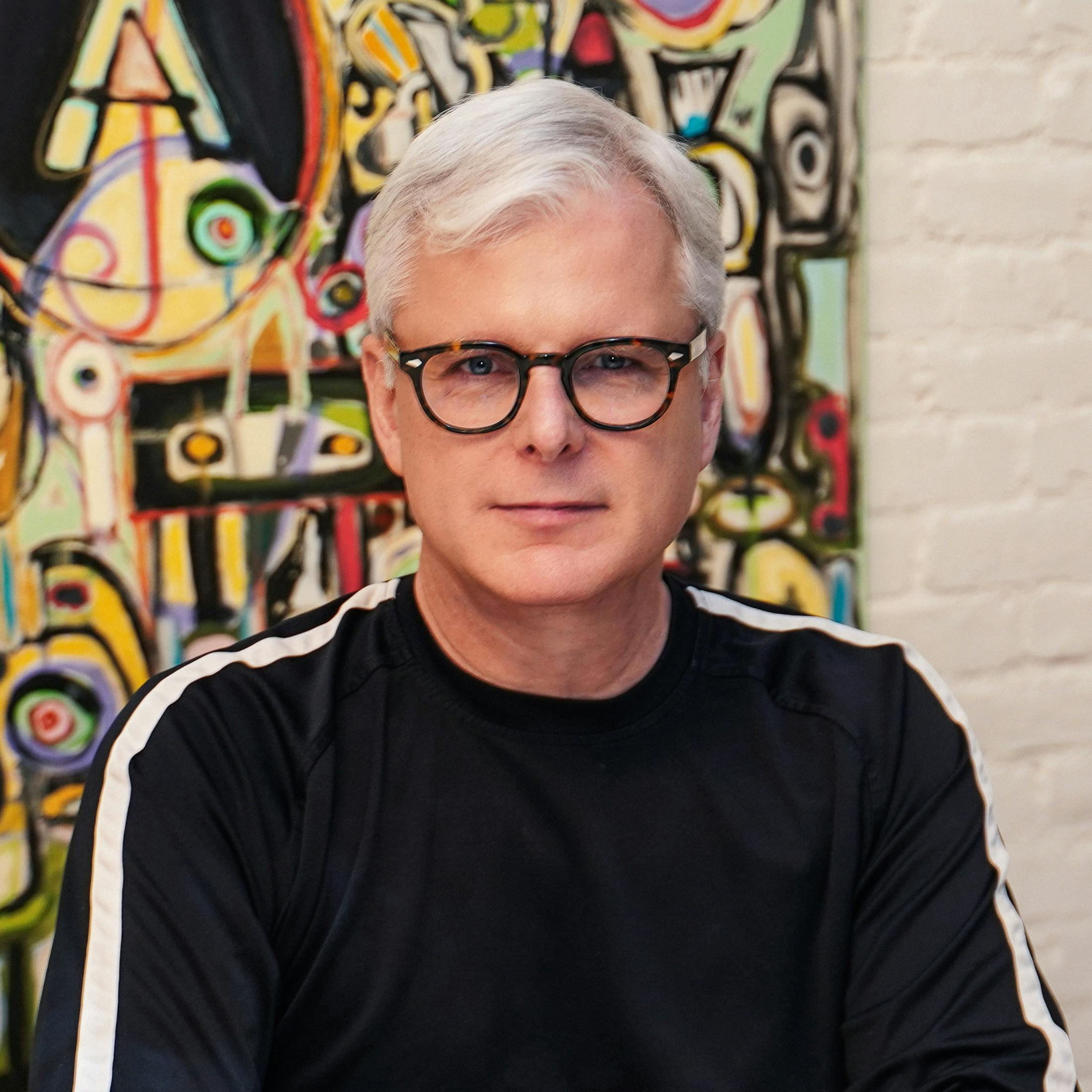 Portrait of StraweberryFrog Head of Strategy Chip Walker wearing tortoiseshell glasses and a black long sleeve with white details in front of an abstract painting. 