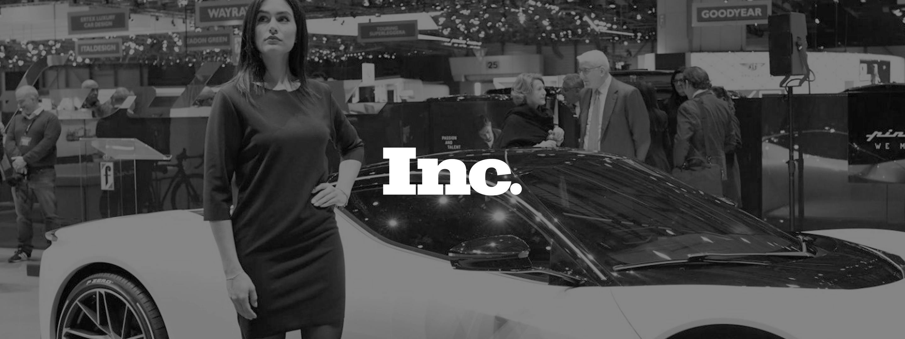 Black and white image of a woman posing next to a concept car with people in the background chatting.